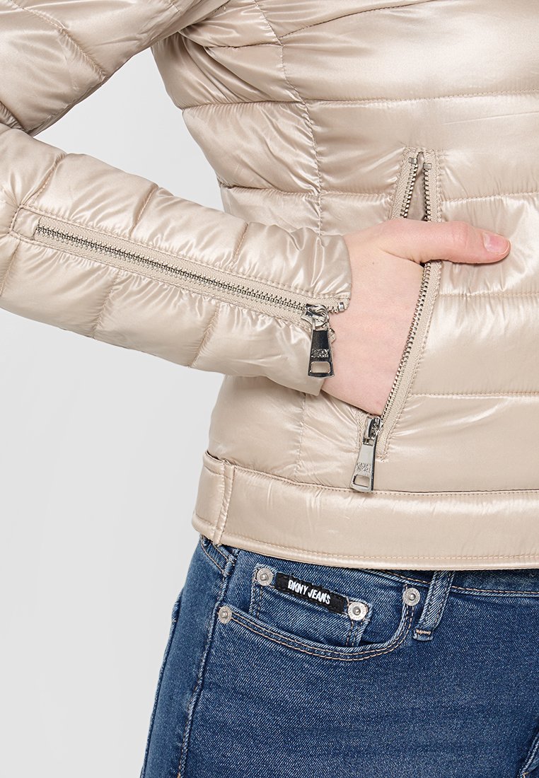 Beige Daunenjacke mit glatter, glänzender Textur, ausgestattet mit Reißverschlusstaschen und horizontalen Nähten. Kombiniert mit blauen Jeans.