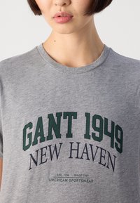 Nærbilde av en person som har på seg en grå t-skjorte med "GANT 1949 NEW HAVEN" og "American Sportswear" trykket foran.