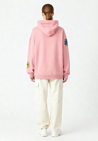 Rosa överdimensionerad hoodie med en framficka, prydd med gula och blå broderade detaljer på ärmarna. Kombinerad med krämfärgade cargobyxor och vita skor.