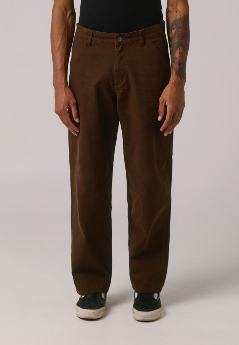 Braune Hosen aus robustem Stoff, mit geradem Bein-Design, Fronttaschen und dezenten Nähten. Getragen mit schwarzen Schuhen.