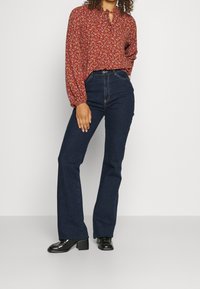 Donna che indossa jeans a vita alta a palazzo blu scuro, scarpe col tacco nere e una camicia a maniche lunghe rossa a fiori, in piedi su uno sfondo neutro.
