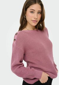 Jeune femme aux cheveux bruns mi-longs portant un pull mauve texturé avec des boutons sur l'épaule et un pantalon noir.