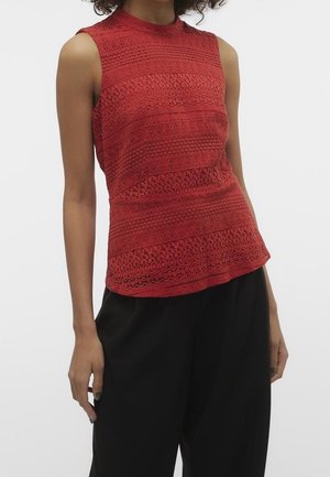 Czerwona koronkowa bluzka bez rękawów, dopasowana, z wysokim kołnierzem, noszona z luźnymi czarnymi spodniami na stojącej osobie na jednolitym tle.