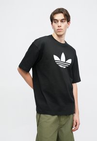 Czarny bawełniany t-shirt z krótkim rękawem i dużym białym logo Adidas z przodu, umieszczony na jednolitym tle.
