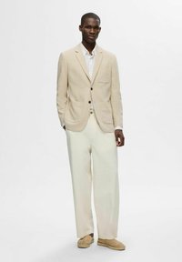 Beżowa marynarka z delikatnym wzorem w kratkę, biała koszula i luźne, kremowe spodnie. Brązowe buty dopełniają stylizację, podkreślając neutralną paletę kolorów.