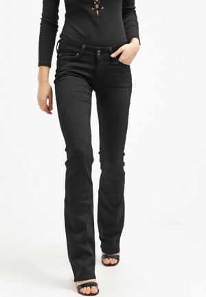 Nainen pukeutuneena mustiin bootcut-farkkuihin, mustaan pitsinauhalla koristeltuun pitkähihaiseen yläosaan ja mustiin avokkaisiin sandaaleihin, kävelee käsi taskussa.