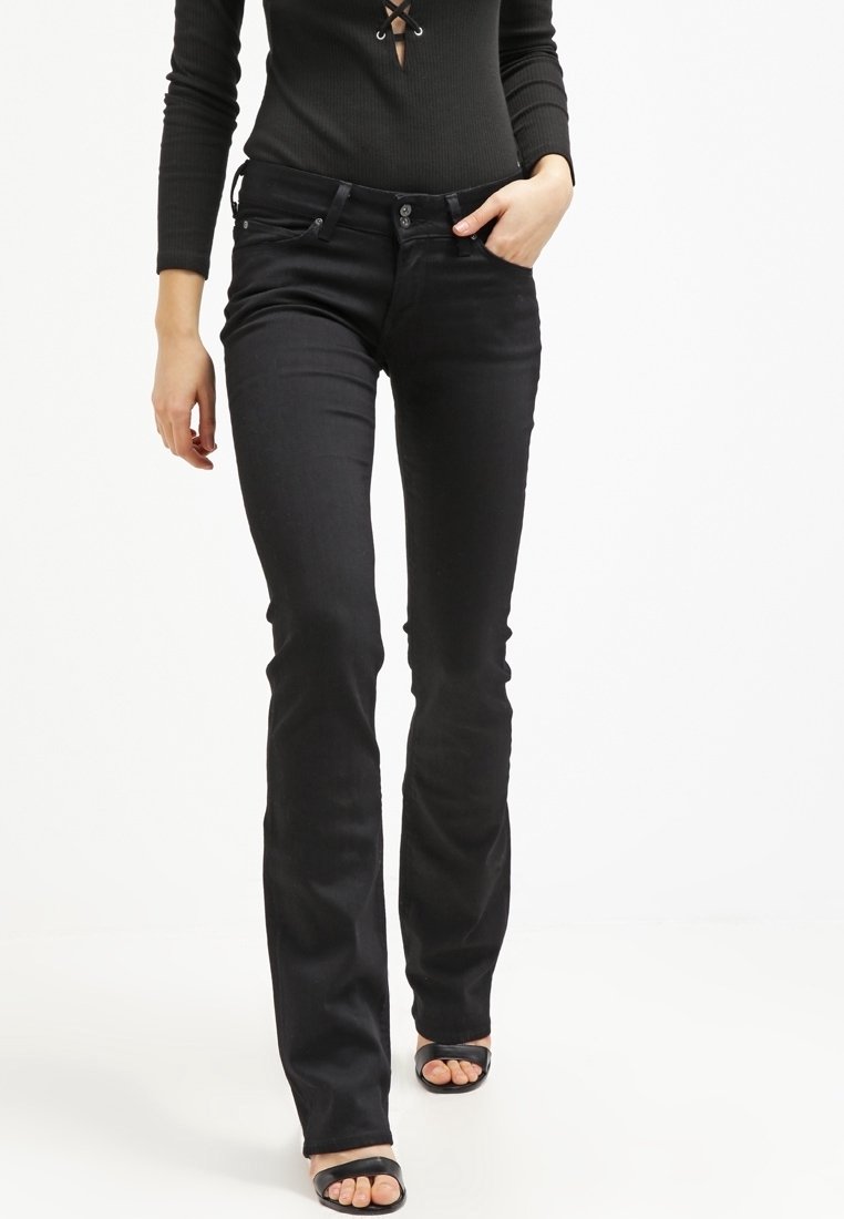 Femme portant un jean noir bootcut, un haut noir à manches longues avec un détail à lacets, et des sandales noires à bouts ouverts, marchant avec une main dans la poche.