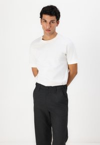 T-shirt blanc à manches courtes, col rond. Pantalon plissé gris foncé avec deux poches avant et deux boutons à la taille. Fond uni.