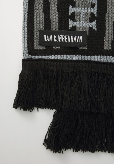 Fekete és szürke kötött sál, bojtokkal a szélein, amelyen egy "HAN KJØBENHAVN" feliratú szövött címke található. A szöveten geometrikus minta látható.
