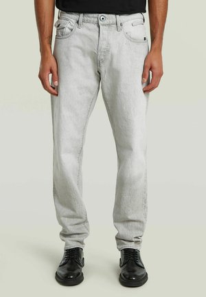 Uomo che indossa jeans grigi chiari a taglio dritto e scarpe eleganti nere con lacci, in piedi contro uno sfondo chiaro e semplice.