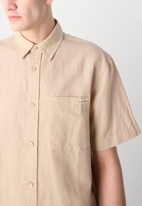 Camicia beige a maniche corte con bottoni, colletto, taschino sul petto e un piccolo logo Calvin Klein. Tessuto di cotone morbido, vestibilità rilassata.