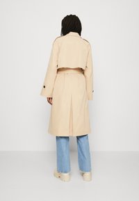 Beige trenčkot s cropped designem, dlouhými rukávy a páskem v pase. Obsahuje zadní rozparek a knoflíkové cuffy. Nosený přes světle modré džíny.