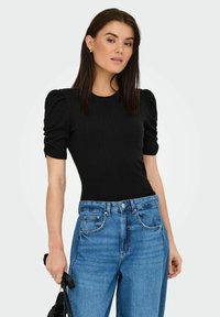 Body in tessuto a coste nero con maniche a sbuffo corte, abbinato a jeans azzurri a vita alta. La modella tiene una borsa nera.