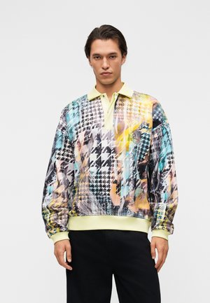 Άνδρας με φαρδιά, μακρυμάνικη πόλο μπλούζα με παστέλ κίτρινο γιακά και πολύχρωμο αφηρημένο σχέδιο houndstooth, σε συνδυασμό με μαύρο παντελόνι.