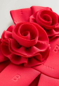 Punane kangast aksessuaar, millel on kihilised rooside detailid selgelt eristuvate kroonlehtedega ning alusosale reljeefne "B" kiri. Sile tekstuur.