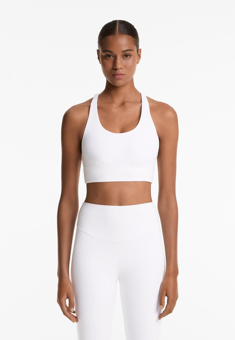 Kobieta z włosami związanymi do tyłu, ubrana w biały sportowy stanik i białe legginsy z wysokim stanem, stoi na tle jednolitego białego tła.