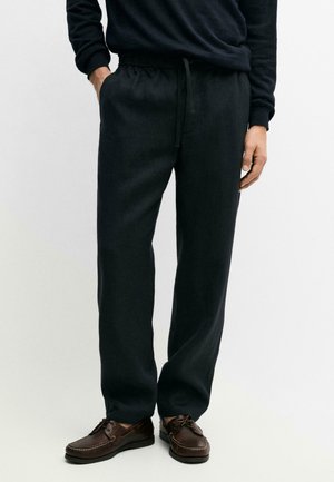 Uomo con pantaloni neri con coulisse e tasche, e scarpe stringate di pelle marrone, in piedi con una mano in tasca.