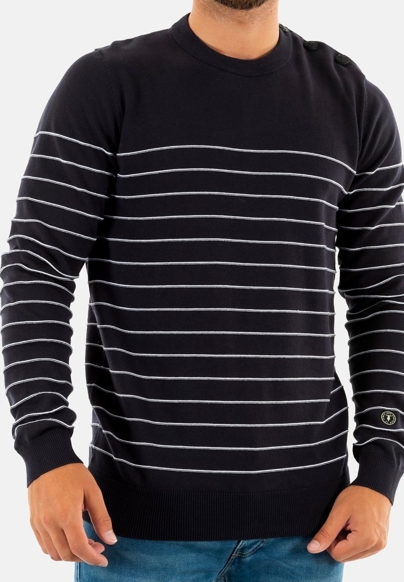 Homme portant un pull noir à manches longues avec de fines rayures horizontales blanches et un jean bleu, debout les mains détendues.