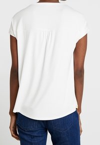Vista trasera de una persona que lleva una blusa blanca de manga corta sencilla con una costura central en la espalda y jeans de mezclilla azul oscuro.