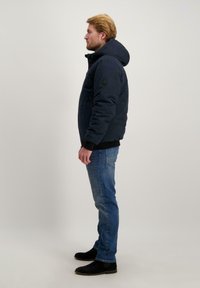 Marineblauwe geïsoleerde jas met capuchon, ritssluiting aan de voorkant en elastische manchetten; gedragen over lichtblauwe jeans en zwarte laarzen.