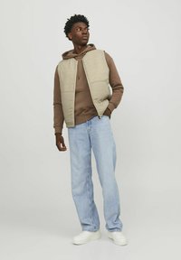 Gilet rembourré beige sur un hoodie marron, associé à un jean droit bleu clair et des baskets blanches. Design simple sans motifs.