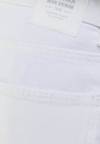 Tela de mezclilla blanca con patrones de costura visibles. La etiqueta dice "MAN DENIM 1998 COLECCIÓN DE MEZCLILLA PREMIUM." Primer plano del área del bolsillo trasero.