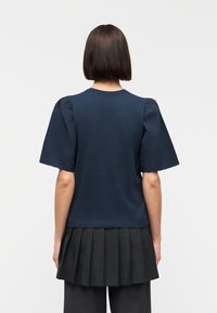 Άτομο με κοντά καστανά μαλλιά που φοράει μπλε μπλούζα με φουσκωτά μανίκια και μαύρη πλισέ φούστα, ορατό από πίσω πάνω σε απλό φόντο.