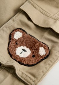 Tissu beige avec un patch en forme d'ours en peluche brun texturé présentant des accents blancs, situé sur une poche à rabat. Une couture noire entoure le patch.