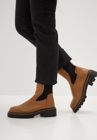 Botas de tobillo de cuero marrón con paneles laterales elásticos, suelas de plataforma negra gruesa y detalles acanalados. Combinadas con jeans negros.