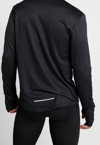 Pitkähihainen musta urheilupaita teksturoidulla kankaalla, jossa on heijastava yksityiskohta takana helman lähellä. Tiimalasimallinen, matala takakappale.