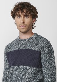 Suéter de punto con cuello redondo que presenta un patrón texturizado en azul claro y oscuro, con una franja horizontal sólida en azul marino a lo largo del pecho.