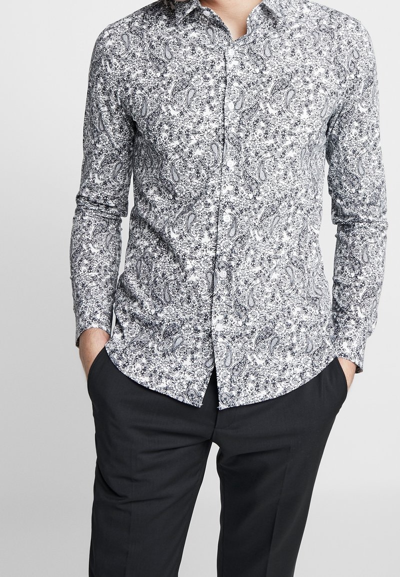 Chemise à motifs paisley noir et blanc avec des manches longues, un col button-down et une coupe ajustée, portée avec un pantalon sombre.