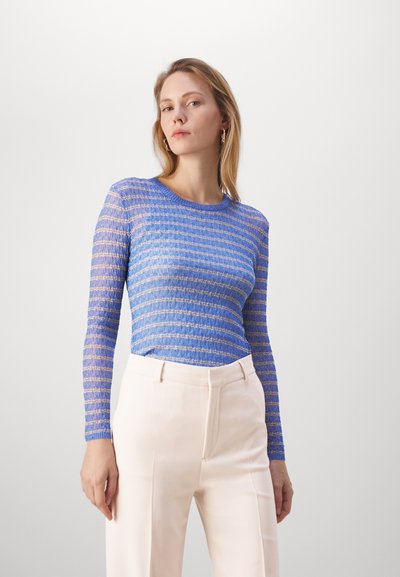 Langärmliges Oberteil in blau und hellblau gestreiftem Strick, mit rundem Hals. Kombiniert mit hellbeigen hochgeschnittenen Hosen.
