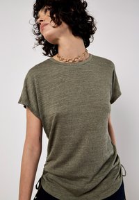 Top de manga corta color verde olivo hecho de tejido texturizado con cordones laterales. Presenta un escote redondo y un corte holgado. Se puede ver un collar de cadena dorada.