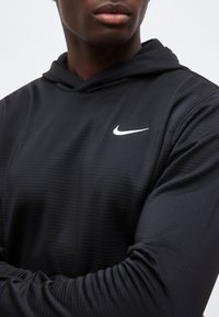 Άνδρας με μαύρο φούτερ Nike με λευκό λογότυπο swoosh, σταυρωμένα χέρια, σε απλό ανοιχτό φόντο.