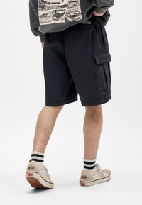 Shorts cargo noirs avec plusieurs poches, coupe décontractée et longueur genou, assortis à des baskets beiges et des chaussettes rayées.