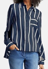 Mujer vistiendo una camisa de botones azul marino con rayas verticales blancas y un bolsillo en el pecho, combinada con jeans azules, con la mano en el bolsillo.