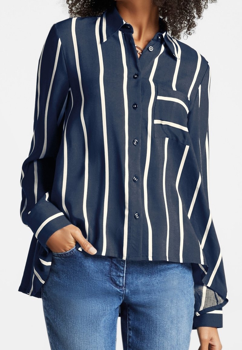 Mujer vistiendo una camisa de botones azul marino con rayas verticales blancas y un bolsillo en el pecho, combinada con jeans azules, con la mano en el bolsillo.