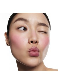 Gros plan d'un visage à la peau claire, avec des joues légèrement rosies, des lèvres brillantes et des cheveux foncés, affichant un clin d'œil espiègle et des lèvres pincées.