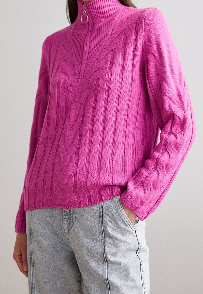 Pull tricoté fuchsia avec motifs en torsades en relief, col montant et détail zippé. Texture côtelée et manches longues. Associé à un denim clair.