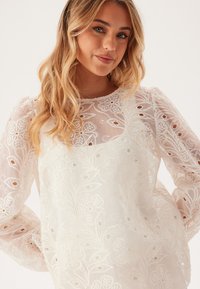 Hvid bluse med gennemsigtig stof, blomsterbroderi og udskæringsdetaljer. Lange ærmer med elastiske manchetter og rund halsudskæring.