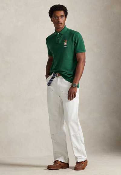 Zelena polo majica z vezenim logotipom, bele hlače sproščenega kroja, rjavi čevlji za čoln in barvit pas. Okrogle očala kot poudarek.
