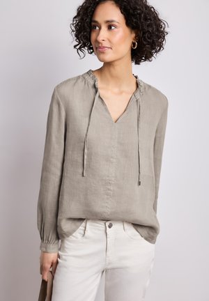 Femme aux cheveux bouclés portant un chemisier ample taupe à manches longues et un pantalon blanc, regardant sur le côté, tenant une pochette.