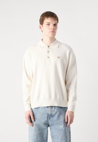 Κρέμ μακρυμάνικο πόλο με γιακά, τρία κουμπιά και μια μικρή ετικέτα λογότυπου στο στήθος. Φορεμένο με ανοιχτό μπλε τζιν.