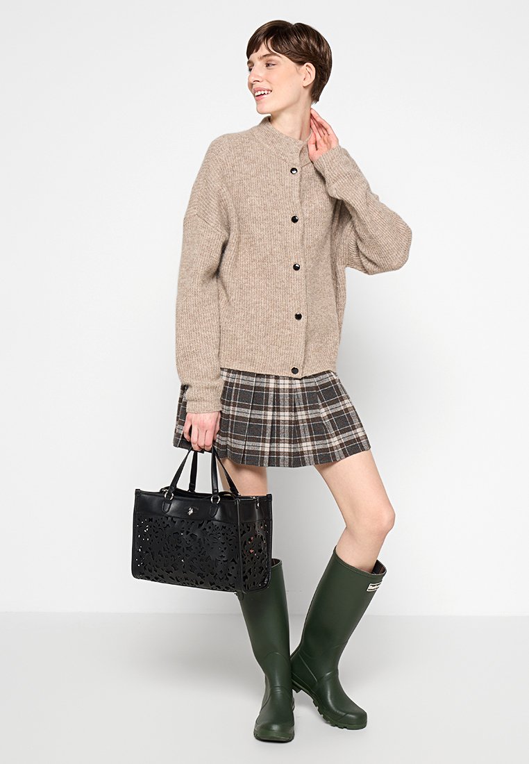 Beige, gestrickte Strickjacke mit Knöpfen, karierten Rock in Brauntönen, schwarze durchlöcherte Handtasche und grüne Gummistiefel, alles vor einem neutralen Hintergrund.