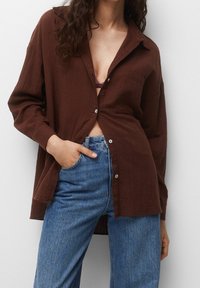 Chemise brune légère à boutons avec un col et une poche avant, portée ouverte sur un haut violet, associée à un jeans bleu clair.