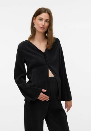 Femme à la peau claire et aux cheveux bruns portant un haut à manches longues plissé noir et un pantalon, touchant doucement son ventre devant un fond uni.
