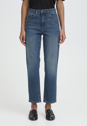 Žena oblečená v modrých džínsoch s rovným strihom, čiernom topu a čiernych mokasínach stojaca pred jednoduchým svetlým pozadím.