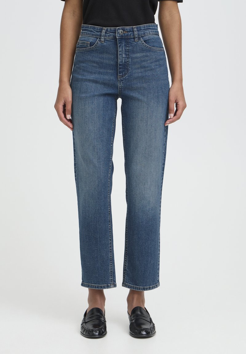 Donna con jeans blu a gamba dritta, maglietta nera e mocassini neri in piedi davanti a uno sfondo chiaro uniforme.