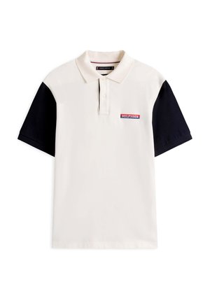 Polo avec un corps crème et des manches courtes noires. Dispose d’une patte de boutonnage et d’un logo brodé rouge, blanc et bleu sur la poitrine.